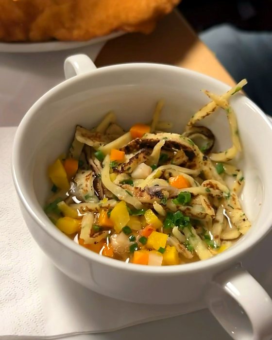 A white bowl of broth with strips of pancake, diced vegetables and chives