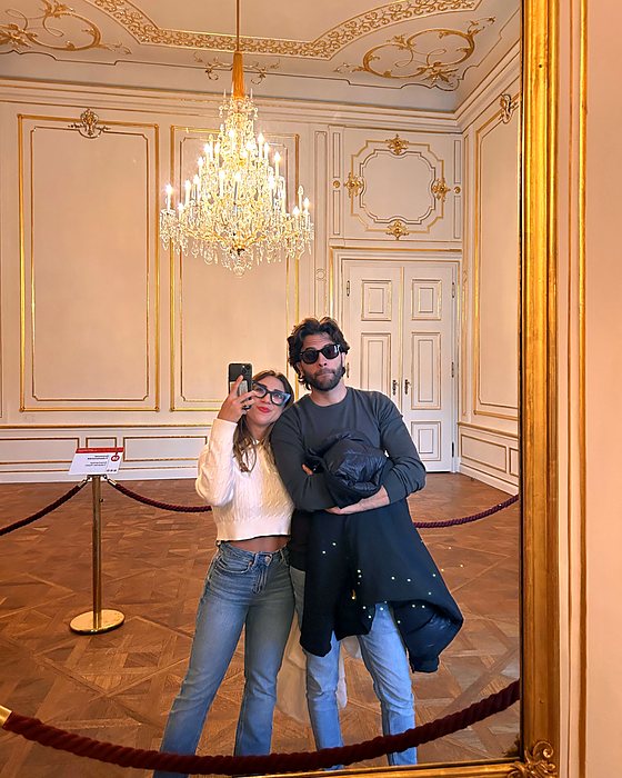 A couple's mirror selfie in a palace room with a large chandelier